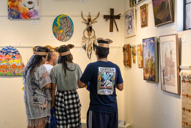 Four fair visitors view paintings, crafts, and mixed-media artworks on display inside an exhibit hall at the New Mexico State Fair.