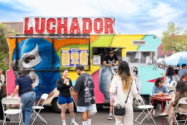 A colorful food truck labeled “Luchador” serves customers as people gather, order food, and sit at nearby tables under a bright sky.