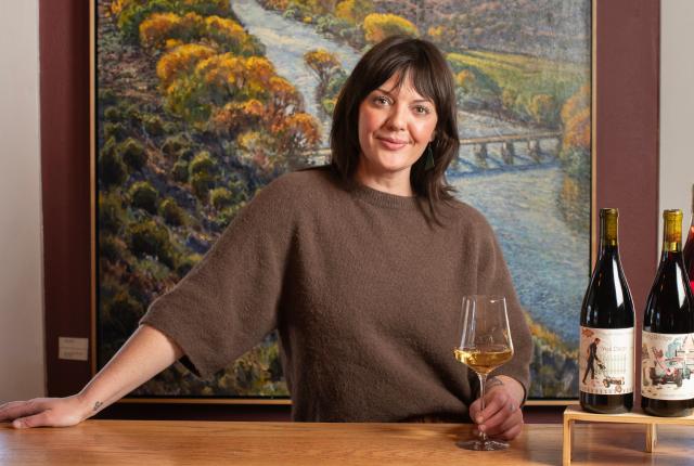A woman winemaker, Katie Hagan, stands behind a bar in the Embudo Valley Vineyards tasting room, pouring wine into a glass.