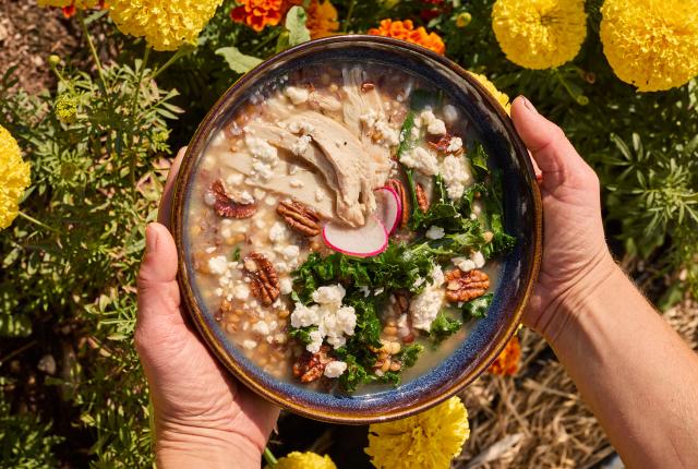 Hands hold a dark bowl filled with grains, greens, and pecans over yellow flowers.