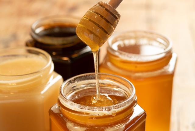 A wooden honey dipper drizzles amber honey into an open glass jar.