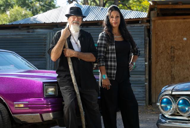An older man with a hat and cane and a woman in a plaid shirt stand between a purple car and a blue classic car, in a sunny setting.