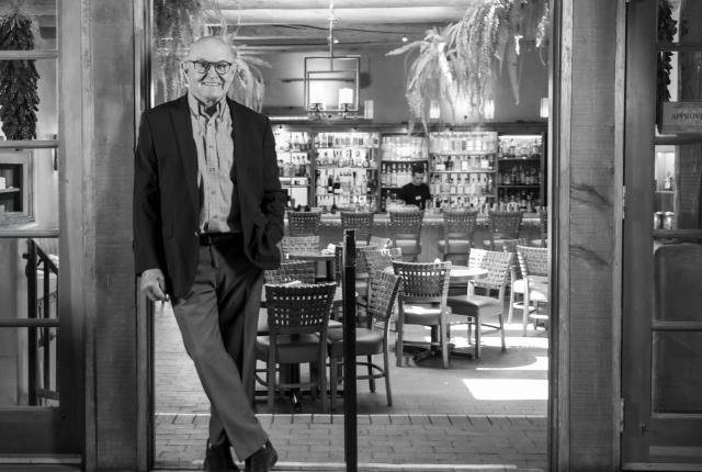A person in a suit stands casually in the doorway of a restaurant decorated with woven patterns, hanging plants, and shelves of bottles in the background.