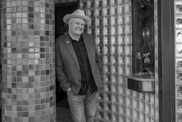 A person wearing a hat and a blazer leans casually against a tiled column outside a glass‑block‑fronted building.