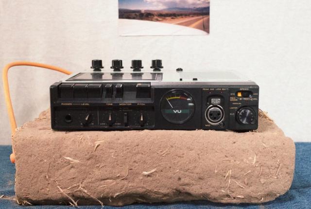 Vintage audio recorder sits on a block of adobe with a landscape photo taped to the wall behind it.