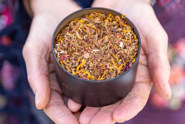 Hands gently holding a black bowl filled with an herbal blend of dried flowers and spices, including yellow petals.