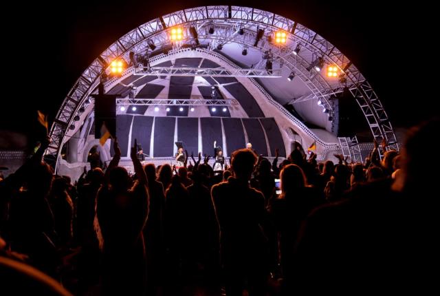 A lively crowd enjoys a nighttime concert under a large, illuminated stage dome.