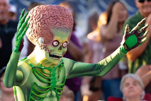 A performer in a bright green alien costume with an oversized textured head and detailed ribbed suit raises both arms while walking through a crowded outdoor event.
