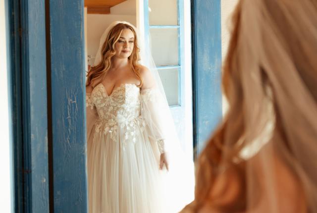 A bride in a lace gown and veil stands near a mirror, framed by a rustic blue door.