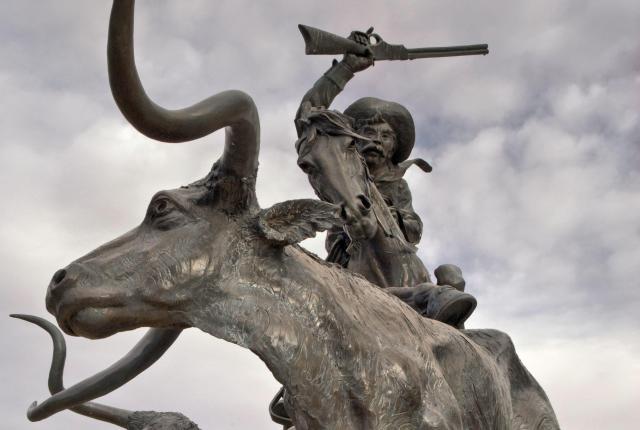 Large bronze sculpture of a rider raising a rifle while leaping forward on a charging longhorn, with additional cattle figures behind.