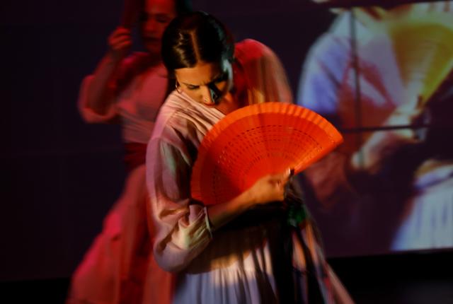 Flamenco dancers on stage holding vibrant orange fans, draped in flowing dresses, move gracefully against a dimly lit backdrop with a large projected image.