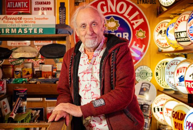 A person stands inside a small vintage‑themed shop surrounded by illuminated retro gas station signs, shelves of old cans, and various memorabilia.