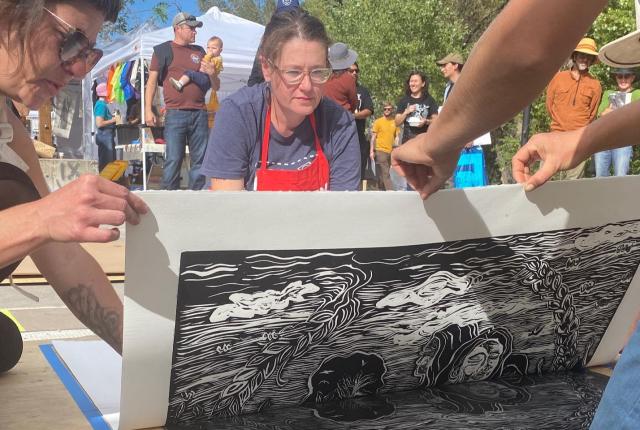 People collaboratively lift a large linocut print from a wooden surface at an outdoor art fair. The black-and-white design features intricate patterns.