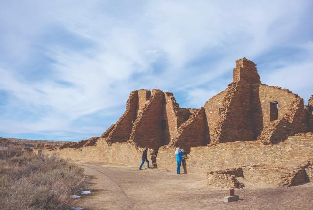 Two people explore ancient stone ruins at Chaco Canyon under a vast, partly cloudy sky.