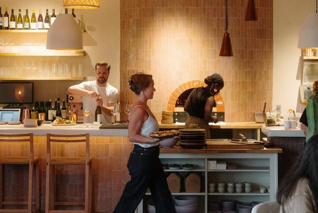 A server carries plates through Tender Fire Kitchen's dining room as cooks work near the stone pizza oven.