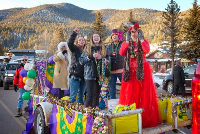 Group of people on a festive Mardi Gras float, wearing bright costumes and masks, celebrating joyfully against a snowy mountain backdrop.
