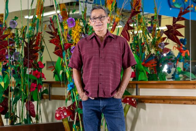 Man in a maroon shirt and jeans stands confidently in a colorful art studio with vibrant plant sculptures and large mirrors.