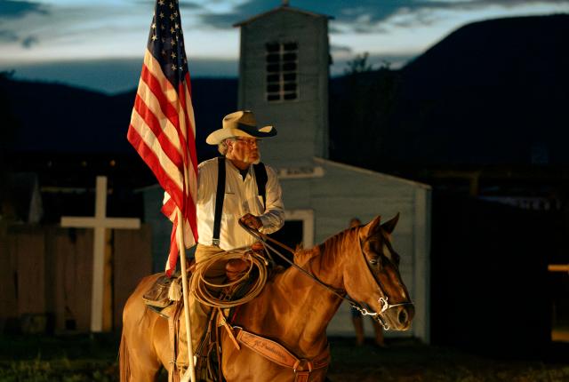 Person riding a horse at dusk while holding an American flag, with a small chapel structure in the background.