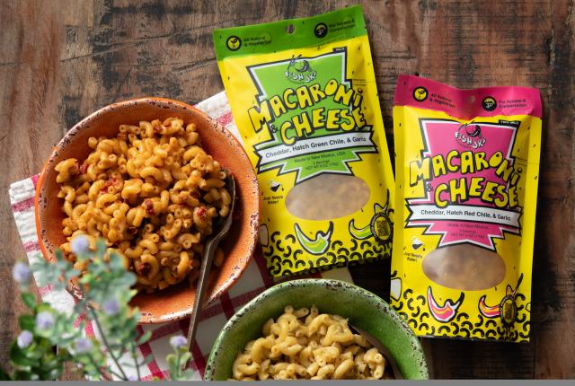 Two colorful bowls of macaroni and cheese, one orange and one green, are placed on a rustic wooden table with vibrant packaging nearby.