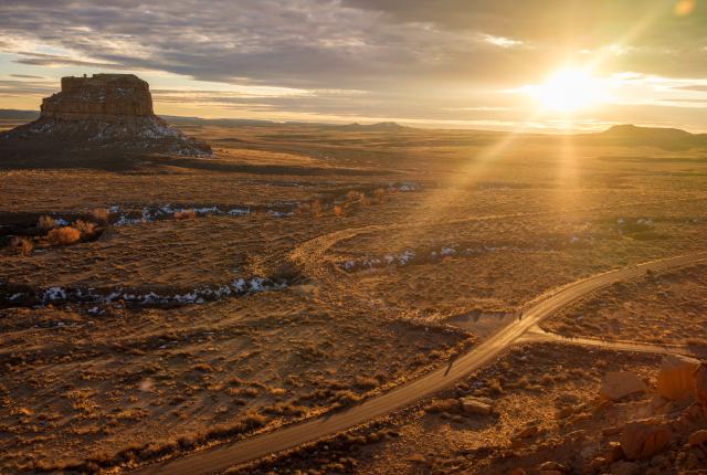 Sunset over a desert landscape with a winding dirt road, a prominent mesa, and scattered brush.