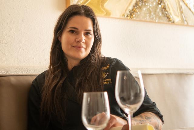A woman with long dark hair, wearing a black chef's jacket, sits at a dining table with empty wine glasses in the foreground.