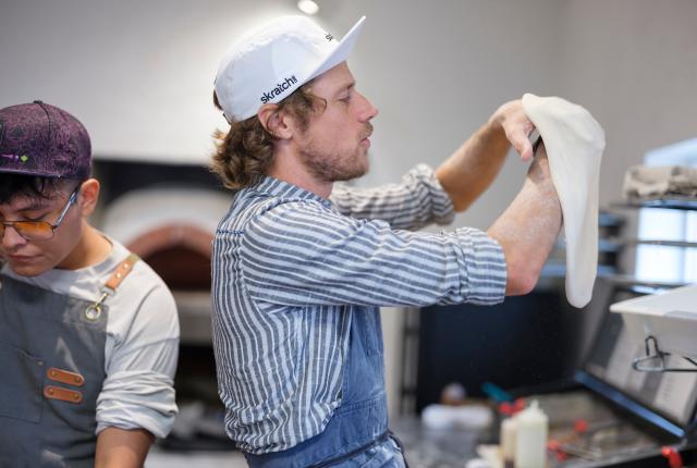 A person in a striped shirt and baseball cap stretches pizza dough in a bright kitchen, while another person in an apron prepares ingredients nearby.