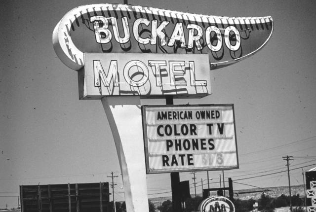 A vintage black‑and‑white roadside sign reads “Buckaroo Motel” with a silhouette of a rider atop a bucking horse above it and additional signage below advertising color TV, phones, and room rates.
