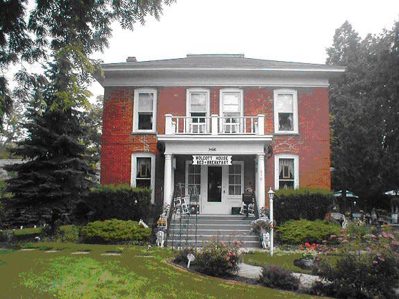 An image of a two story red brick home surrounded by a pine tree on the left and other trees around the home. Home has white balcony with below leading to the front door.