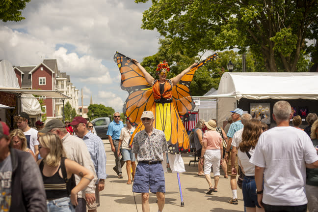 A stilt walker dressed as a monarch butterfly moves through a crowded outdoor arts festival lined with vendor tents.