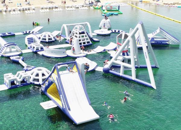 Image of water park located in Anaheim, California. Image shows inflatable obstacles placed inside a large pool of water with people swimming and climbing on each obstacle.