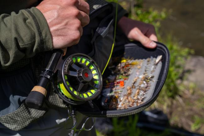 A close-up of a person holding a fly fishing reel and a tackle box filled with various flies, set against a natural backdrop.