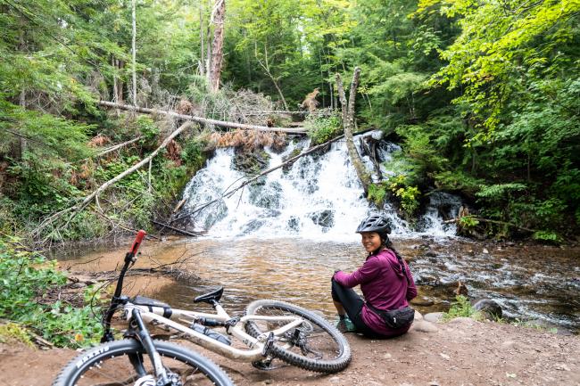 A woman sitting next to her bike in front of a waterfall, smiling in Marquette, MI