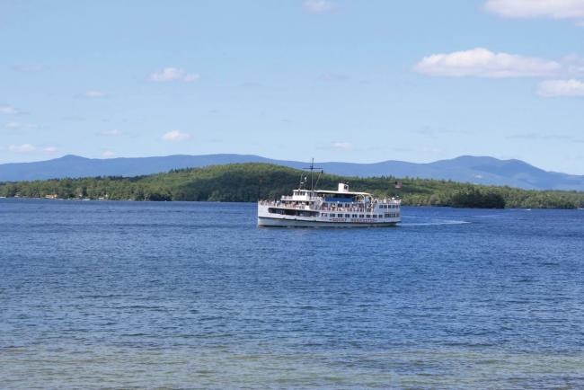 A scenic view of the Mount Washington cruise ship on a lake, with lush green hills and distant mountains under a blue sky.
