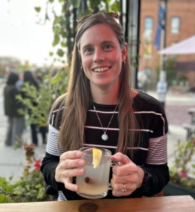 Woman sits outdoors at a table and smiles for the camera hlding a beverage in a glass cup in her hands.