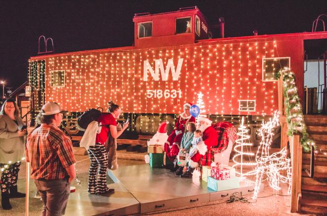 A red NW train car draped in lights stands behind a photo op with Santa