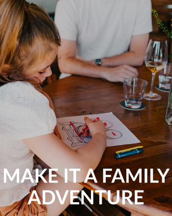 A child coloring at a table inside a winery tasting room, with adults and wine glasses in the background.