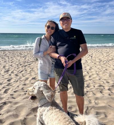 Man and woman link arms and smile for a photo while standing on sand at the beach with ocean water in the background. A dog lays in front of the couple in the sand.