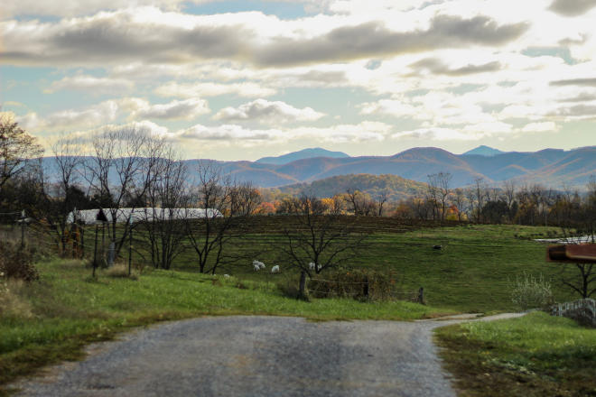 Sunlight illuminates Blue Ridge Mountains' fall color through the clouds. A gravel road and farm sits in the foreground.