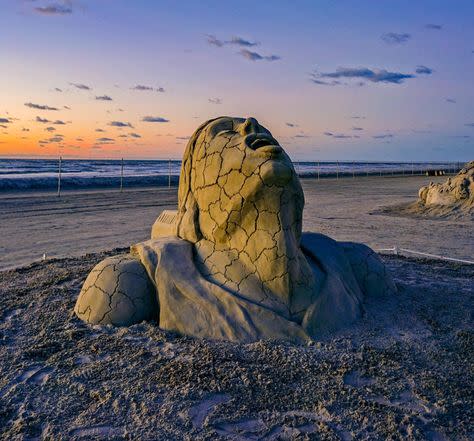 Sand sculpture of a face looking up to the sky, mouth agape, with cracks running through the face.