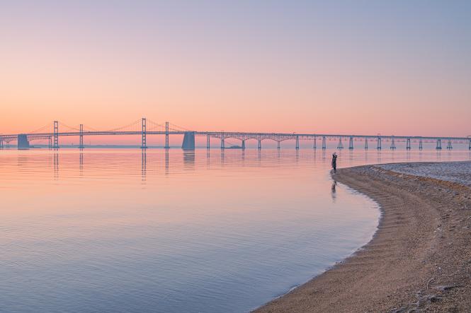 A peaceful calm bay with a bridge in the background and a woman standing at the edge of the water during the winter