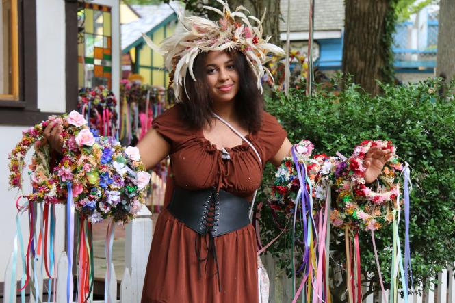A woman dressed in medieval attire holds flower crowns on her arms