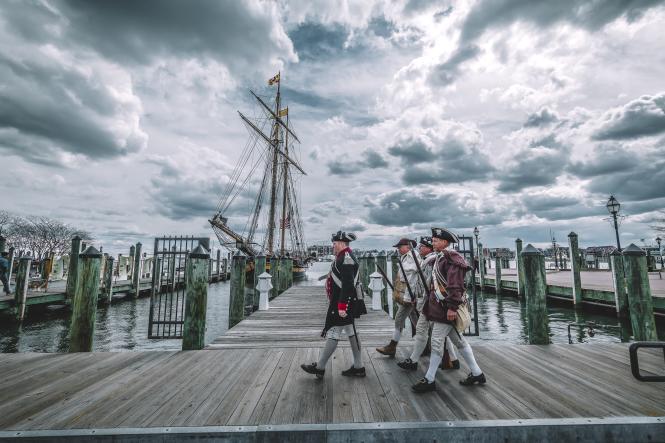 Men in period military costumes walking on a dock past Tall Ships in a harbor