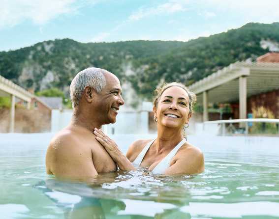 A couple smiles in a hot spring with green hillsides in the background in Glenwood Springs