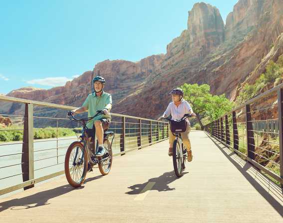 Two people bike on a path with towering red rock formations in the background in Moab, Utah
