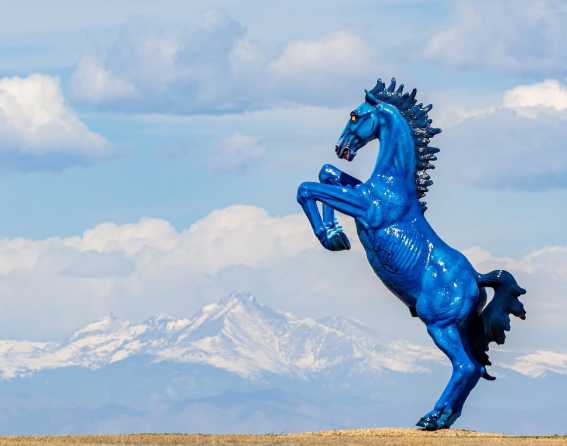 Blucifer is shown on a blue-sky day with snowcapped Rocky Mountains in the background