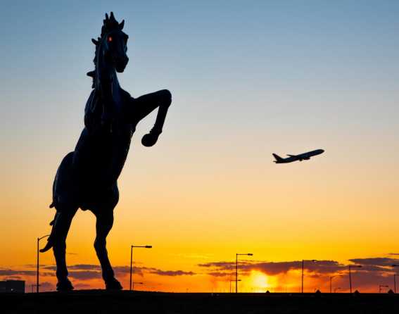 A silhouette of Blucifer is shown at sunset with a plane taking off in the background