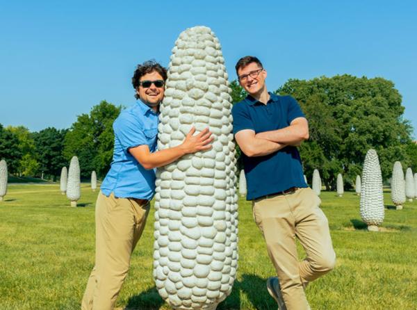 A couple of friends at the Field of Corn. One of them hugging a corn and the other leaning on a corn.