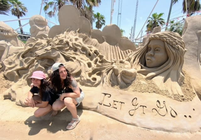 Two women kneel next to a sand sculptor that shows a woman blowing something out of her cupped hands. it looks to be a vague horse. There's a banner under her that reads, "Let It Go"
