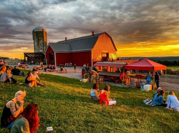 a sunset in back of a red barn with people sitting on the lawn in the foreground