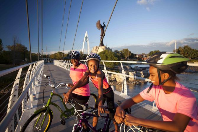 A mom and two middle school age children park their bikes on a pedestrian bridge overlooking the Keeper of the Plains